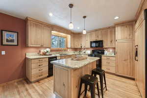 Kitchen featuring hanging light fixtures, light stone counters, black appliances, light hardwood / wood-style floors, and a kitchen island