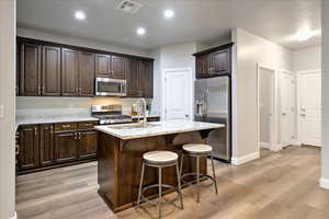 Kitchen featuring a kitchen island with sink, light stone counters, stainless steel appliances, and dark brown cabinetry