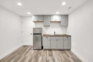 Kitchen with gray cabinetry, sink, stainless steel refrigerator, and butcher block counters. Location in recreation/living room in the basement.