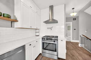 Kitchen with white cabinetry, wall chimney range hood, decorative light fixtures, and stainless steel appliances. Pantry set just behind this wall.