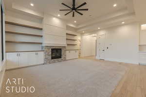 Unfurnished living room featuring a tray ceiling, a stone fireplace, ceiling fan, and light wood-type flooring