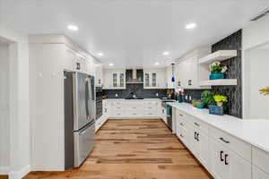 Kitchen featuring wall chimney range hood, white cabinetry, stainless steel appliances, decorative backsplash, and light wood-type flooring