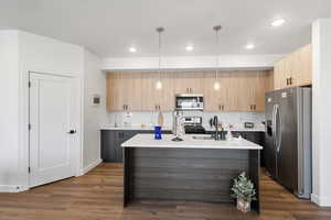 Kitchen with dark wood-type flooring, appliances with stainless steel finishes, a center island with sink, light brown cabinetry, and decorative light fixtures