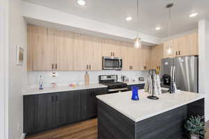 Kitchen with dark hardwood / wood-style flooring, hanging light fixtures, a kitchen island with sink, stainless steel appliances, and light brown cabinets