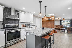 Kitchen with white cabinetry, a kitchen breakfast bar, hanging light fixtures, stainless steel gas range oven, and wall chimney range hood