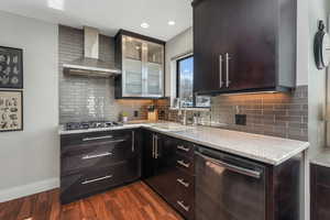 Kitchen featuring tasteful backsplash, stainless steel appliances, light stone countertops, dark wood-type flooring, and wall chimney range hood