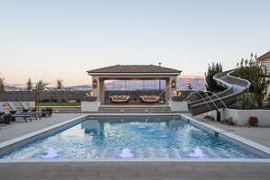 Pool at dusk featuring a water slide, a gazebo, outdoor lounge area, a patio, and pool water feature