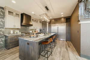 Kitchen featuring range, a kitchen island with sink, premium range hood, light stone counters, and stainless steel built in fridge