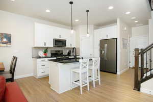 Kitchen featuring light wood-type flooring, appliances with stainless steel finishes, pendant lighting, a kitchen island with sink, and white cabinets