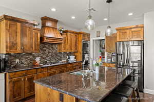 Kitchen featuring sink, a center island with sink, custom range hood, pendant lighting, and stainless steel appliances