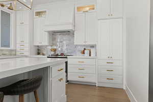 Kitchen with a breakfast bar area, premium range hood, white cabinetry, decorative backsplash, and light wood-type flooring