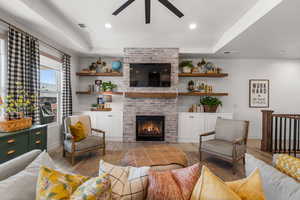 Living room with ceiling fan, a raised ceiling, a brick fireplace, and light wood-type flooring