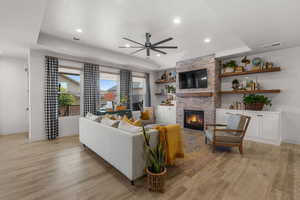 Living room with a brick fireplace, a raised ceiling, ceiling fan, and light wood-type flooring