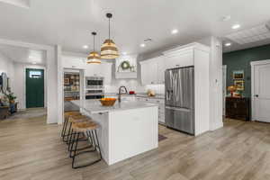 Kitchen with a kitchen island with sink, hanging light fixtures, stainless steel appliances, white cabinets, and light wood-type flooring