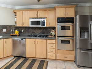 Kitchen with light brown cabinetry, decorative backsplash, ornamental molding, light tile patterned floors, and stainless steel appliances