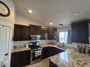 Kitchen featuring stainless steel appliances, vaulted ceiling, dark brown cabinetry, and light stone countertops