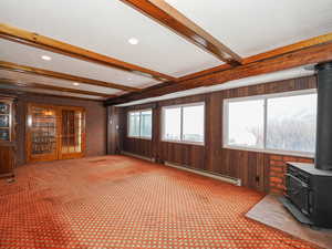 Unfurnished living room featuring light carpet, beamed ceiling, baseboard heating, and a wood stove