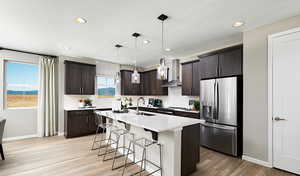 Kitchen featuring wall chimney exhaust hood, dark brown cabinets, a center island with sink, appliances with stainless steel finishes, and pendant lighting