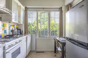 Kitchen featuring wall chimney range hood, white appliances, light tile patterned floors, white cabinets, and decorative backsplash