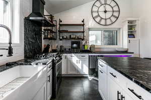 Kitchen featuring vaulted ceiling, white cabinetry, dark stone countertops, range with two ovens, and wall chimney range hood