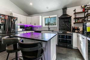 Kitchen featuring vaulted ceiling, white cabinetry, a kitchen breakfast bar, black appliances, and wall chimney range hood