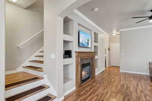 Living room featuring hardwood / wood-style flooring, crown molding, ceiling fan, a tiled fireplace, and built in shelves