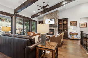 Living room with wood-type flooring, a stone fireplace, vaulted ceiling with beams, and ceiling fan