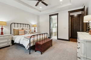 Bedroom featuring ceiling fan, ensuite bath, a tray ceiling, and light carpet