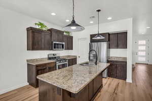 Kitchen featuring appliances with stainless steel finishes, a kitchen island with sink, dark brown cabinets, light stone countertops, and light wood-type flooring