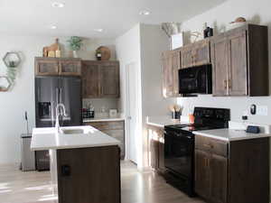 Kitchen with sink, dark brown cabinets, light wood-type flooring, a center island with sink, and black appliances