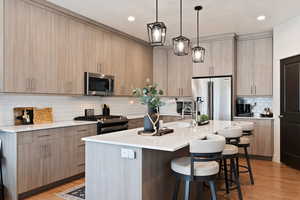 Kitchen with light hardwood / wood-style flooring, hanging light fixtures, stainless steel appliances, a center island with sink, and light brown cabinets