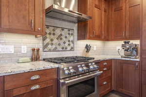 Kitchen featuring stainless steel gas range oven, exhaust hood, backsplash, and light stone counters