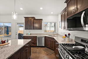 Kitchen featuring dark brown cabinetry, sink, light stone counters, hanging light fixtures, and stainless steel appliances