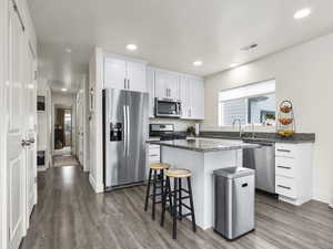 Kitchen featuring dark wood-type flooring, appliances with stainless steel finishes, a center island, white cabinets, and dark stone counters