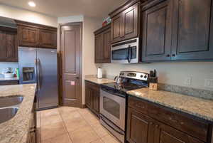 Kitchen featuring dark brown cabinetry, light stone countertops, light tile patterned floors, and stainless steel appliances