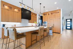 Kitchen with pendant lighting, range hood, a kitchen breakfast bar, light hardwood / wood-style floors, and a large island with sink