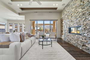 Living room with a stone fireplace, built in features, wood-type flooring, ceiling fan, and a tray ceiling