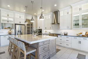 Kitchen featuring wall chimney exhaust hood, decorative light fixtures, appliances with stainless steel finishes, an island with sink, and white cabinets