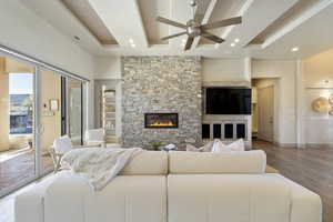 Living room with ceiling fan, wood-type flooring, a stone fireplace, and a tray ceiling