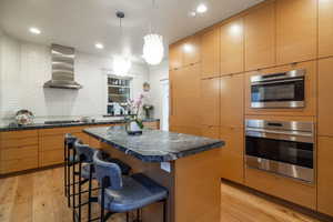 Kitchen with decorative light fixtures, a kitchen island, wall chimney range hood, light hardwood / wood-style floors, and backsplash