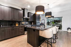 Kitchen with wall chimney exhaust hood, dark brown cabinetry, decorative light fixtures, stainless steel appliances, and a kitchen island with sink