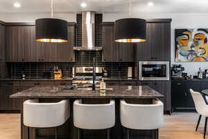 Kitchen featuring wall chimney exhaust hood, a kitchen bar, a kitchen island with sink, and dark stone countertops