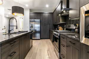 Kitchen featuring wall chimney exhaust hood, sink, dark brown cabinets, dark stone counters, and stainless steel appliances