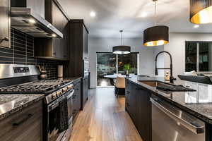 Kitchen with decorative light fixtures, sink, dark stone counters, stainless steel appliances, and wall chimney range hood