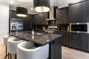 Kitchen with wall chimney exhaust hood, stainless steel appliances, a kitchen island with sink, and hanging light fixtures