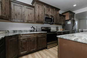 Kitchen with appliances with stainless steel finishes, dark brown cabinets, light stone counters, a textured ceiling, and light wood-type flooring