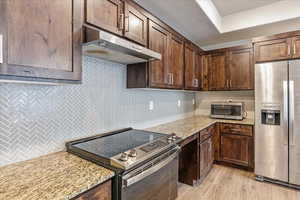 Kitchen with appliances with stainless steel finishes, tasteful backsplash, dark brown cabinetry, light stone counters, and light wood-type flooring