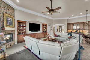 Living room with ornamental molding, a stone fireplace, built in features, and ceiling fan