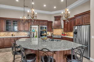 Kitchen featuring appliances with stainless steel finishes, a kitchen breakfast bar, custom range hood, an island with sink, and decorative light fixtures