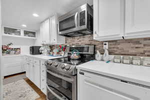 Kitchen with wood finished floors, white cabinets, appliances with stainless steel finishes, decorative backsplash, and glass insert cabinets